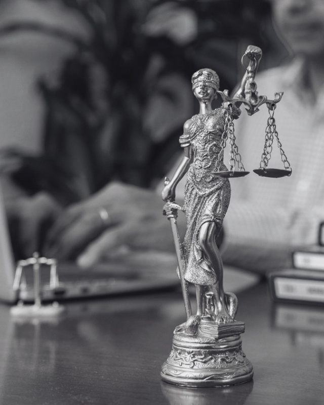 Estatuilla de la justicia sobre el escritorio, con Andrea trabajando en el computador al fondo.