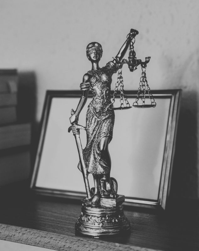 Estatuilla de la justicia con la balanza, sobre el escritorio, delante de un portarretratos y una pila de libros.